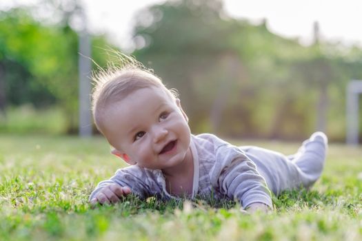 Cute funny laughing Caucasian baby boy learning to crawl, having fun playing on the lawn watching summer flowers in the garden during bright sunny day. Photo of smiling small 6 months old boy in green grass - he smiles and has fun while looking up. Happy young baby lying on tummy in nature.